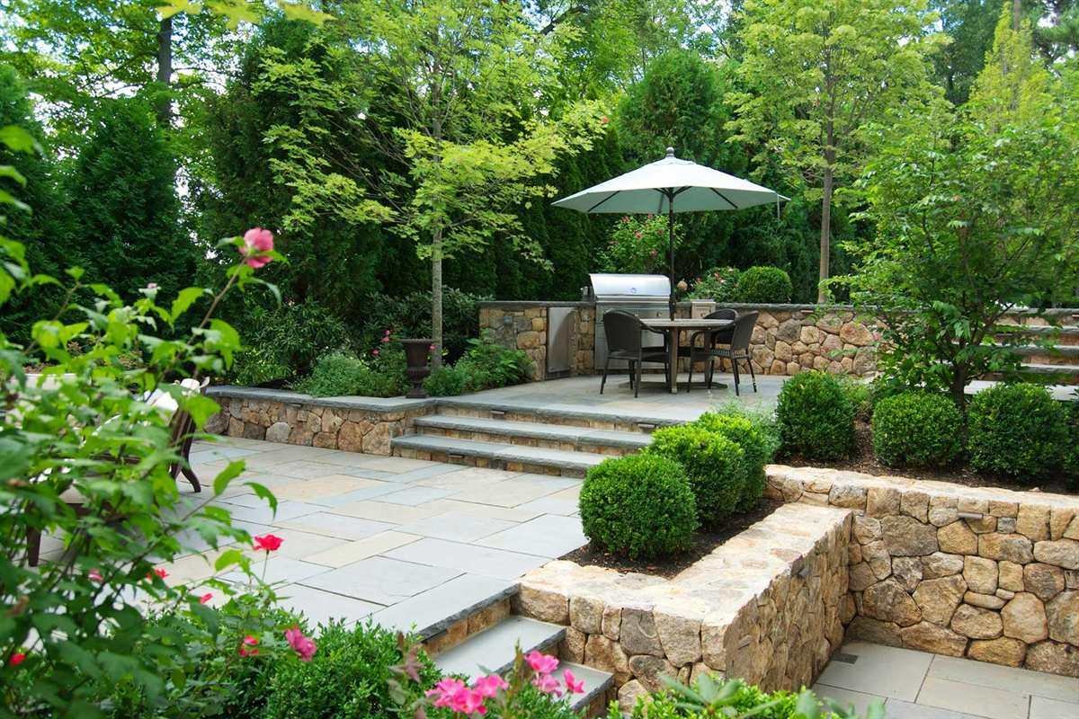 Raised stone terrace with stone steps, a dining set, boxwood plantings and a low garden wall
